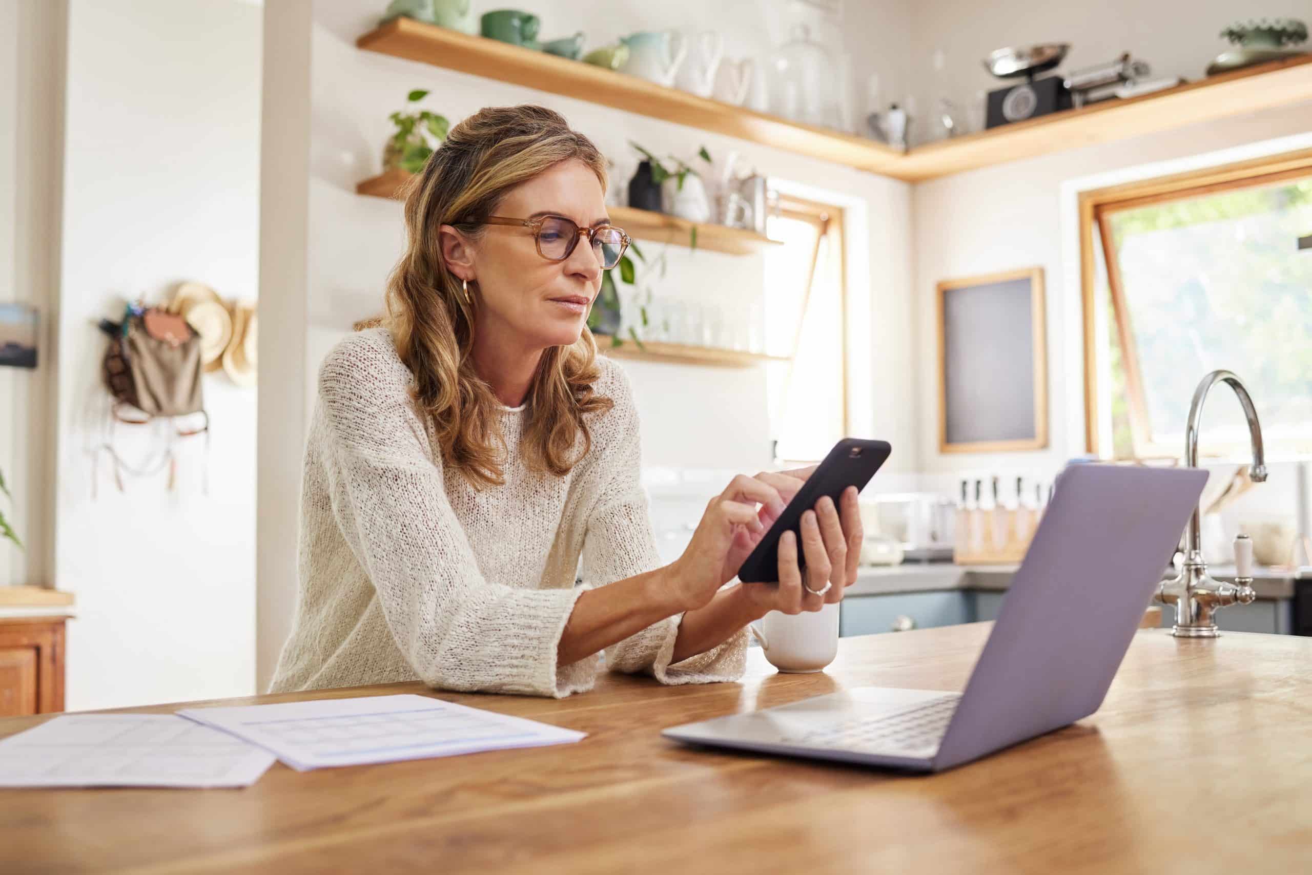 Frau mit Laptop und auf dem Smartphone schauend zu Hause in der Küche, um den Energieverbrauch Ihrer Neubauwohnung in Berlin zu verfolgen