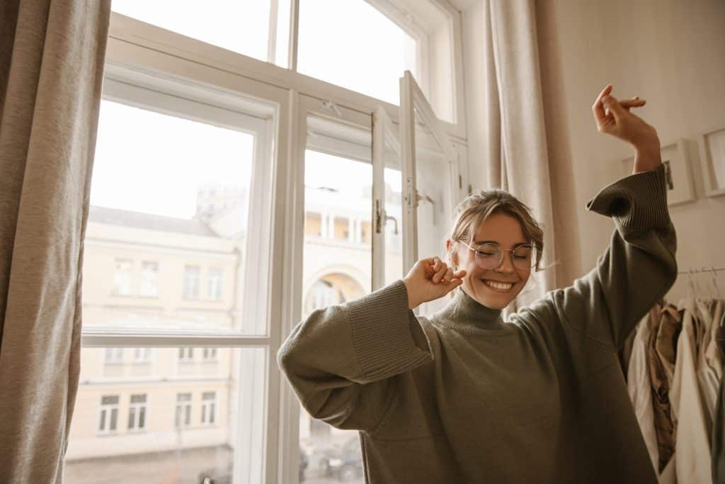 Jeune femme souriante dansant les yeux fermés, seule chez elle en journée. Photo évoquant la joie après l'achat d'un appartement à Berlin
