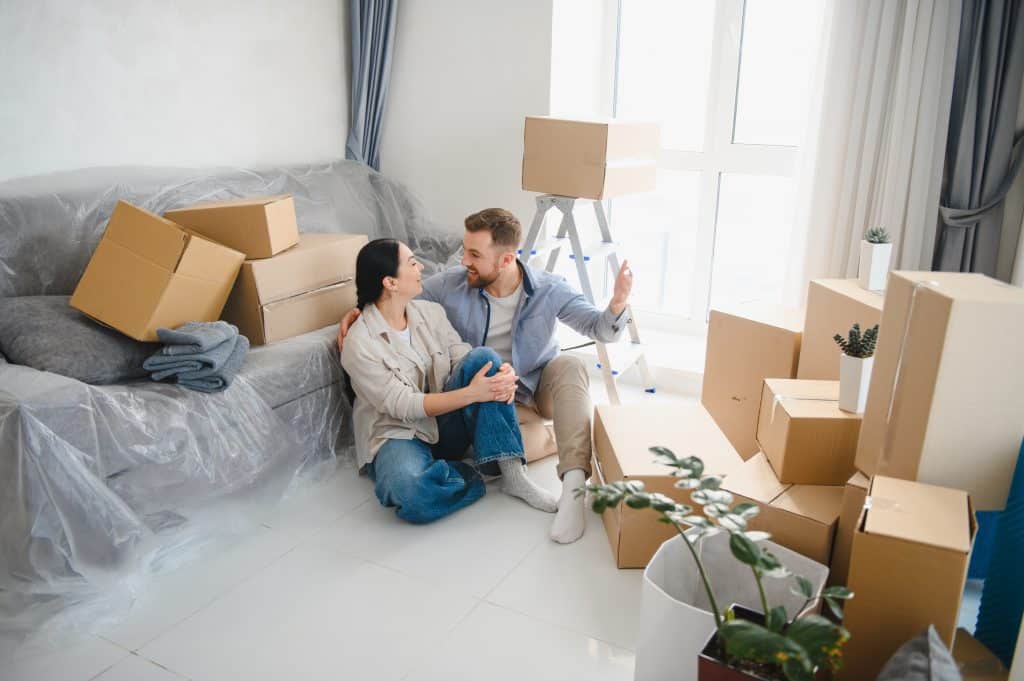 Ein junges Paar sitzt in seiner neuen Wohnung auf dem Boden, umgeben von Kartons, und feiert den Umzug und die Zukunft